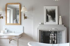 a neutral vintage bathroom with dove grey walls, a grey tub, a fireplace clad with tiles, a mother of pearl chandelier and a free-standing tub