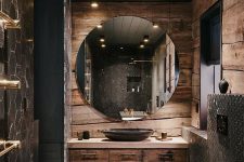 a refined moody bathroom clad with rough wood, with a black planked wooden ceiling, a round mirror, a tiled floor and matching walls