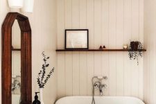 a small and cozy bathroom clad with white planks, a small tub, a vanity with a stone countertop, a mirror in a wooden frame