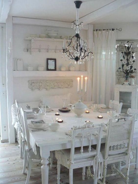 a white shabby chic dining room with lace linens, mismatched chairs, a crystal chandelier and open shelving