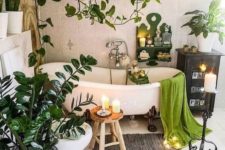 a boho chic bathroom with a vintage tub, some lights and candles, potted plants all around the tub and suspended