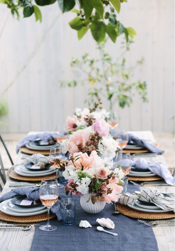 a bright spring table setting with navy napkins and a table runner, wicker chargers and blue porcelain and pink and white floral centerpieces
