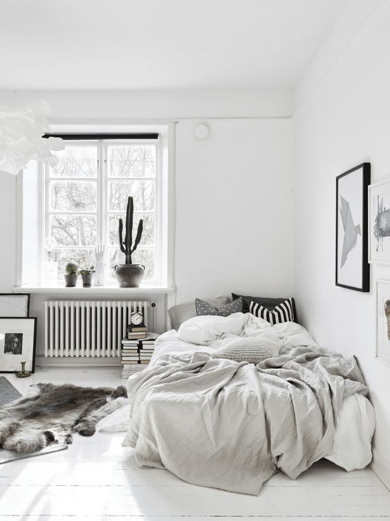 a monochromatic Nordic bedroom with some potted cacti, a bed in the corner, books and artworks