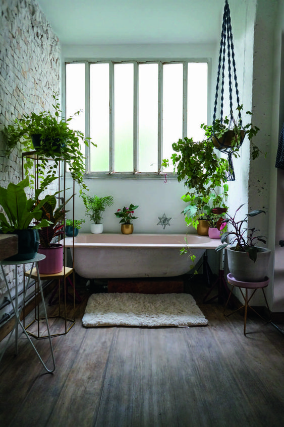 a pretty moody bathroom with a pink tub, colorful planters with greenery all around and a brick wall for interest