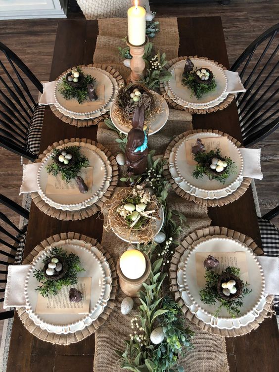 a rustic Easter table setting with a burlap runner and placemats, white porcelain, nests with eggs, greenery, candles is a cool idea for a party