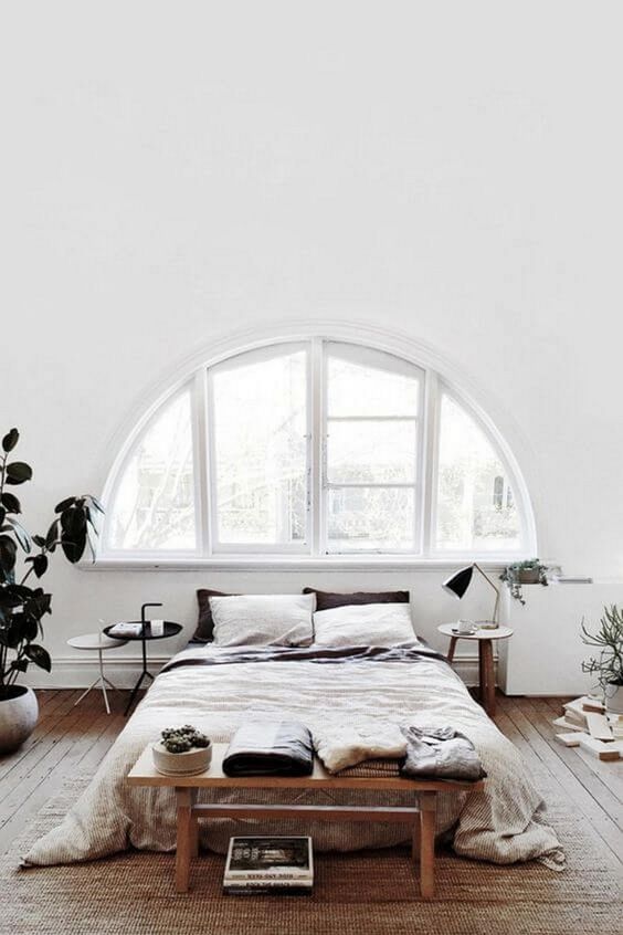 an airy white Nordic bedroom with an arched window, a bed, some wooden furniture and potted greenery
