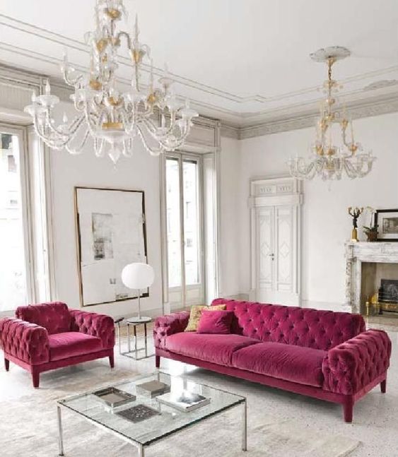 a chic vintage-inspired interior with a faux fireplace, a fuchsia sofa and chair, a glass coffee table and a duo of crystal chandeliers