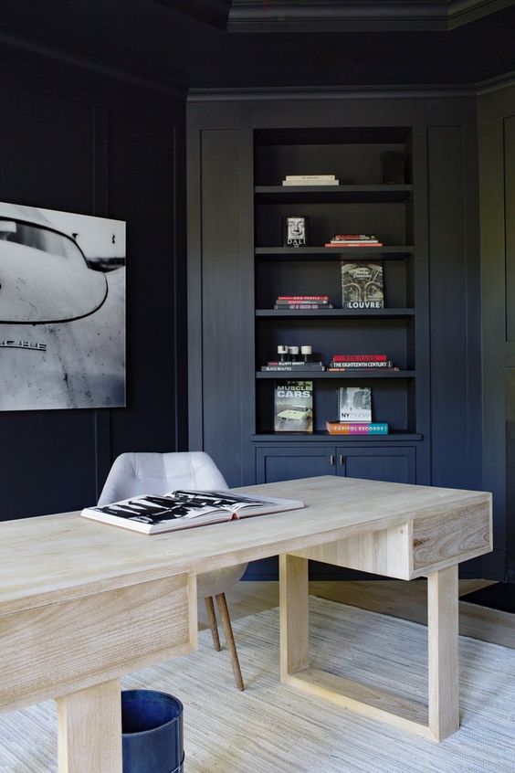A contrasting home office with navy walls and built in furniture, a bleached wooden desk and a statement artwork on the wall