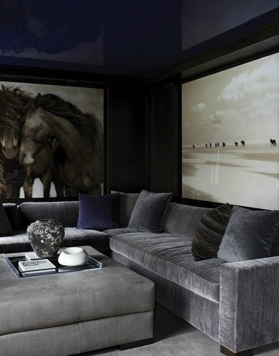 a dark living room with grey furniture, oversized artworks, an upholstered ottoman and a rug