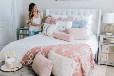 a modern feminine bedroom with a white upholstered bed, mirror nightstands, a grey bench, pink and white bedding and floral artworks