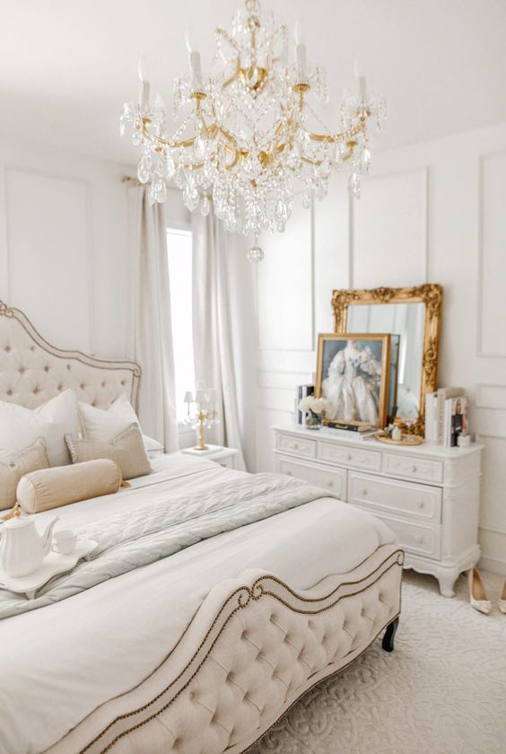 A neutral Versaille inspired bedroom with refined furniture, a crystal chandelier, gold framed art and mirrors and layered bedding
