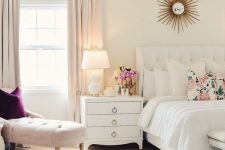 a neutral feminine bedroom with a white upholstered bed, white nightstands, a blush couch and a purple pillow and blush curtains