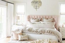 a relaxed feminine bedroom with wooden beams, a pink upholstered bed, a leather bench, neutral bedding and a chic chandelier