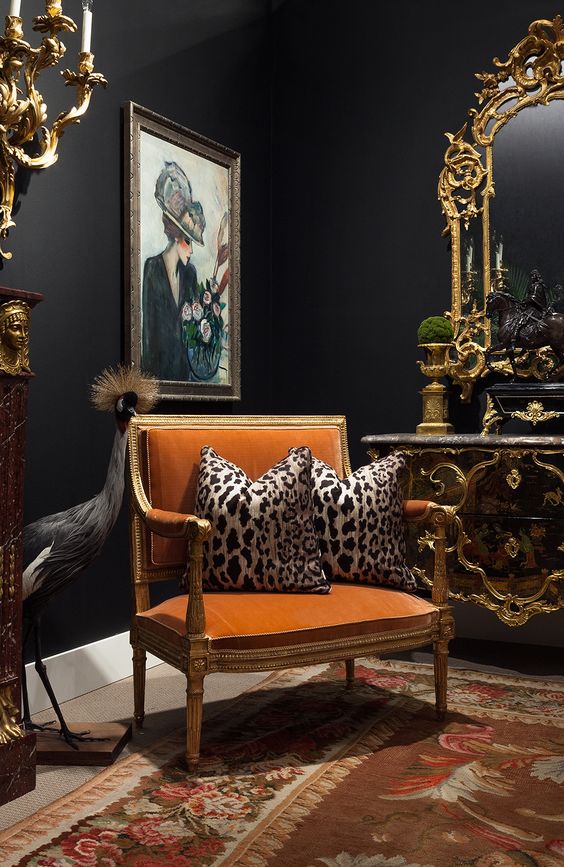 a sophisticated moody living room with black walls, a printed rug, a vintage vanity with a mirror in a gilded frame over it, an orange chair and leopard print pillows