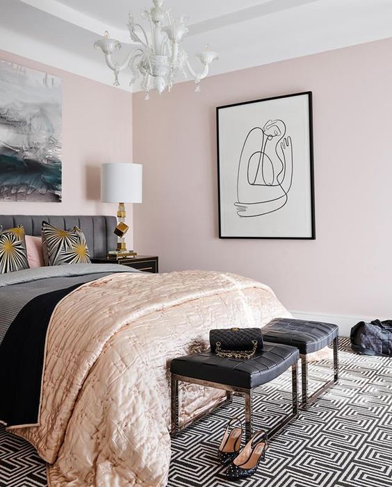 a super stylish feminine bedroom with light pink walls, a grey bed, black woven stools, a statement artwork and touches of gold