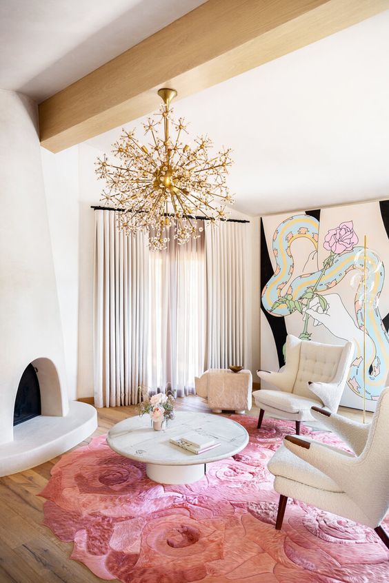 a unique feminine living room with a fireplace, a unique artwork on the wall, neutral chairs, a pink rose statement rug and a gold chandelier