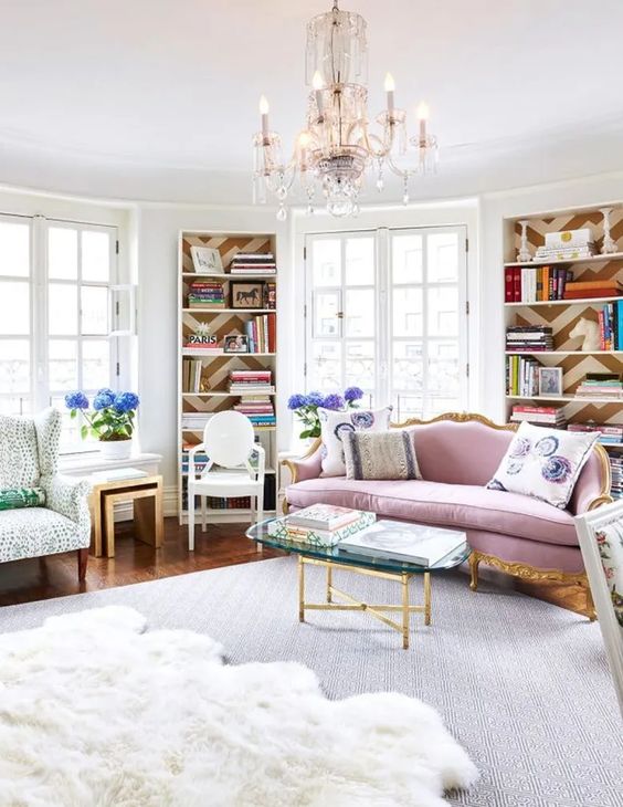 an elegant and refined feminine living room with a chic lilac sofa, bookcases, a printed rug and chairs, a glass coffee table and a crystal chandelier