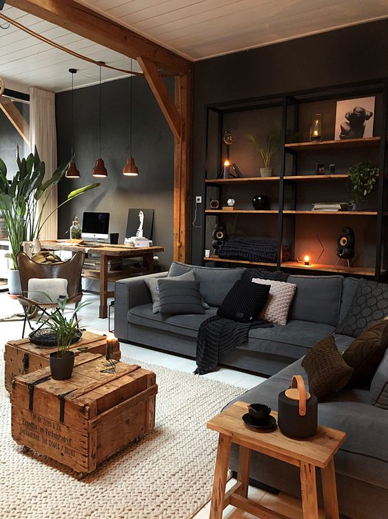 An industrial living room with a wall mounted shelving unit, grey upholstery, wooden chests and a stool and workspace in the corner