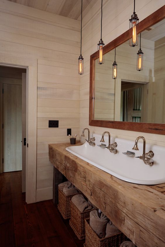 a barn bathroom clad with whitewashed wooden planks, with a rough wooden vanity, pendant lamps and baskets for storage