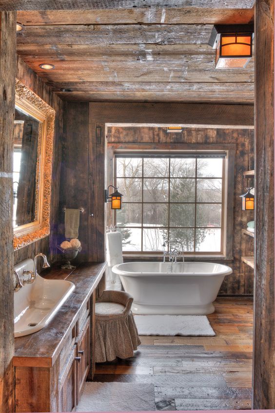 A barn bathroom decorated with rough weathered wood, with a large window, a wooden vanity and a free standing tub