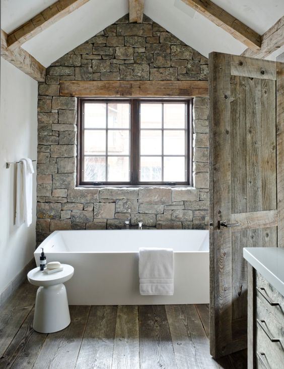 A barn bathroom with a stone wall, a wooden floor and wooden beams, a free standing tub, a window for natural light