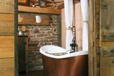 a barn bathroom with a stone wall, wood plank walls, a metal tub and vintage chandeliers plus a jute rug