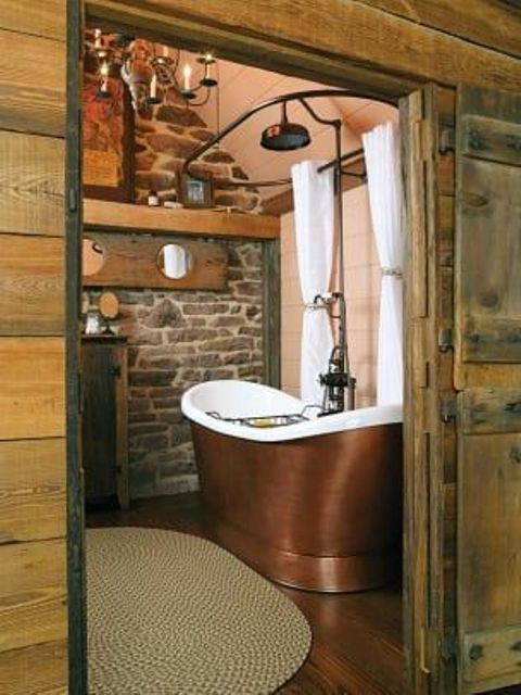 a barn bathroom with a stone wall, wood plank walls, a metal tub and vintage chandeliers plus a jute rug