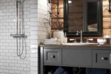 a barn bathroom with a wooden wall, a vintage grey vanity, a white tile clad shower space and a mirror in a black frame