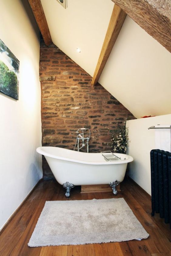 a barn bathroom with white walls and a ceiling, wooden beams, a stone wall, a clawfoot tub and an artwork
