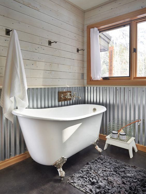 a barn bathroom with whitewashed wooden planks, corrugated steel on the walls, a clawfoot tub, a large window and a fluffy rug