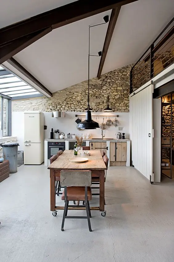 a barn kitchen with a stone accent wall, a glazed one, planked cabinets, dark wooden beams and a vintage dining set of wood and metal