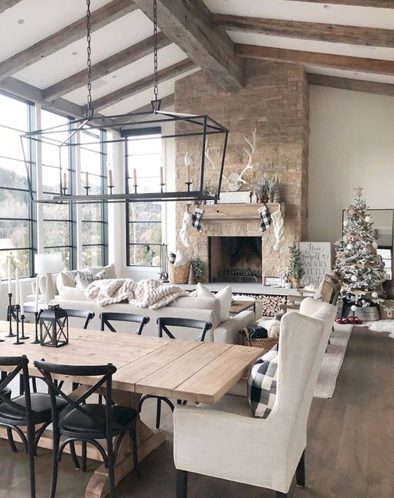 a barn living room with a glazed wall, a fireplace clad with stone, wooden beams on the ceiling, neutral seating furniture and pendant lamps