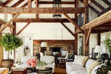 a barn living room with multiple wooden beams, a fireplace clad with stone, white seating furniture, a stained coffee table and potted greenery