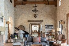 a barn living room with stone walls, wooden beams on the ceiling, brown, grey and white furniture, a piano, refined metal chandeliers and low coffee tables