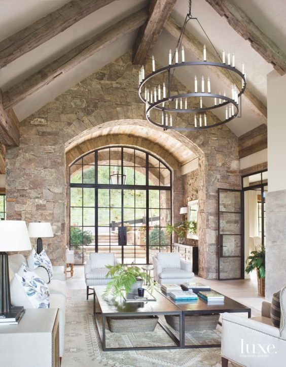 a barn living room with stone walls, wooden beams on the ceiling, neutral seating furniture, low coffee tables and a metal chandelier