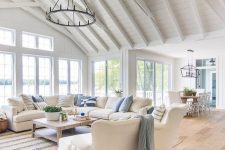 a beach barn living room in white, with woodne beams, white seatign furniture, printed textiles, a metal round chandelier, lovely views of the beach