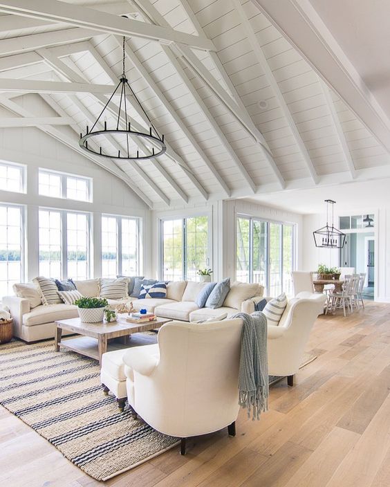 a beach barn living room in white, with woodne beams, white seatign furniture, printed textiles, a metal round chandelier, lovely views of the beach