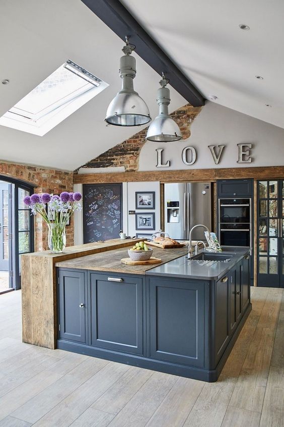 a beautiful barn kitchen with a skylight, a navy wooden beam with lamps, slate grey cabinets and a kitchen island with reclaimed wood