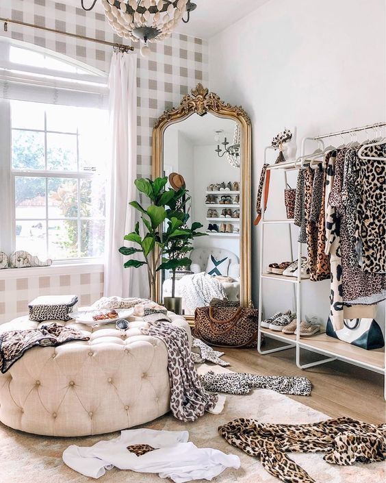 a bold feminine closet with an open storage unit, a shoe shelf, a mirror in an ornated frame, a neutral tufted pouf and a potted plant