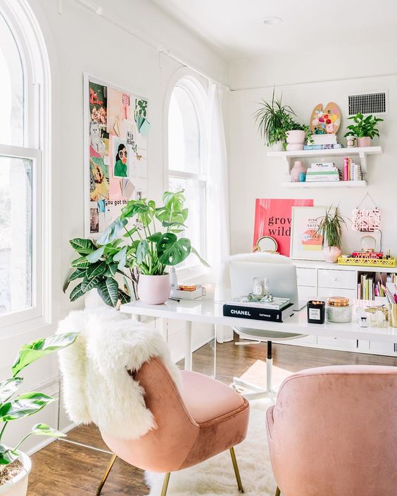 a bright home office with blush chairs, a colorful gallery wall, bright artworks and books and lots of greenery