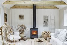 a coastal barn living room in white, with wooden beams, a metal hearth, white seating furniture, reclaimed wood items and dried blooms