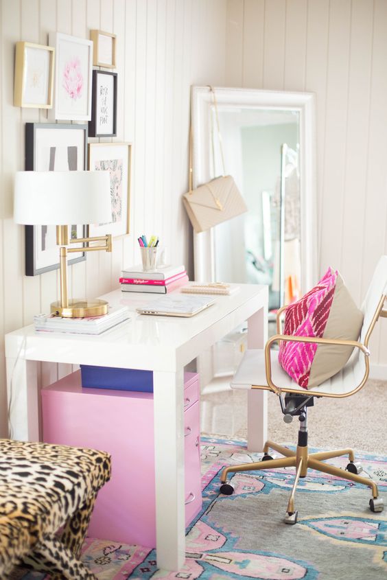 a colorful feminine home office with a white desk, a pink cabinet and pillow, a bright rug and a chic gallery wall