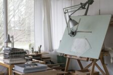 a cool and simple home artist studio with a large window, a wooden table and a table on casters, some shelves with supplies and a large easel