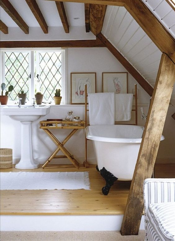 a cozy barn bathroom with white walls and a ceiling, stained wooden beams, a clawfoot tub and vintage furniture