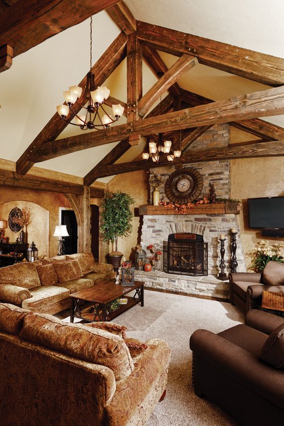 a cozy barn living room with neutral walls, wooden beams, a fireplace clad with stone, velvet and leather brown furniure and pendant chandeliers