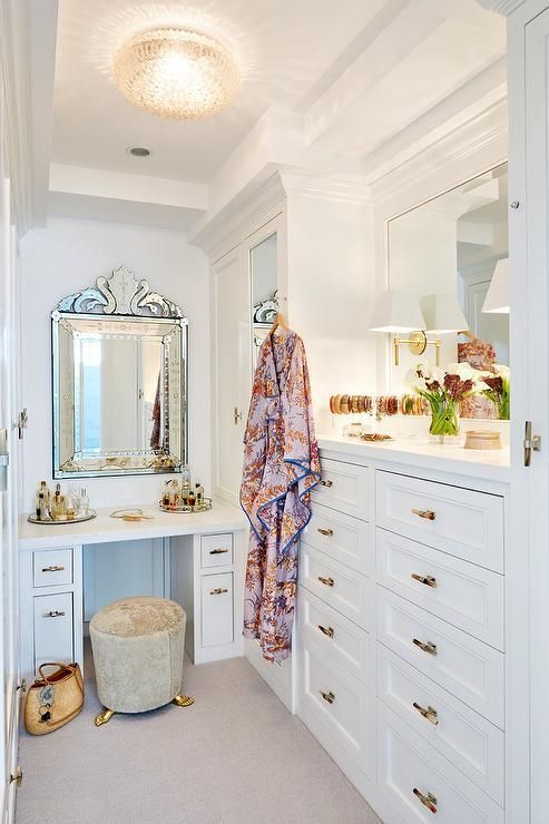 a delicate and chic small walk-in closet with white closed storage units and drawers, a white vnaity, a couple of mirrors and touches of gold