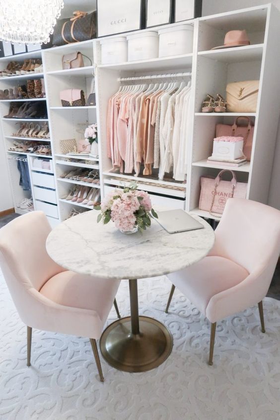 a delicate and subtle feminine closet with open shelves and storage units, pink chairs and a round stone table plus a crystal chandelier