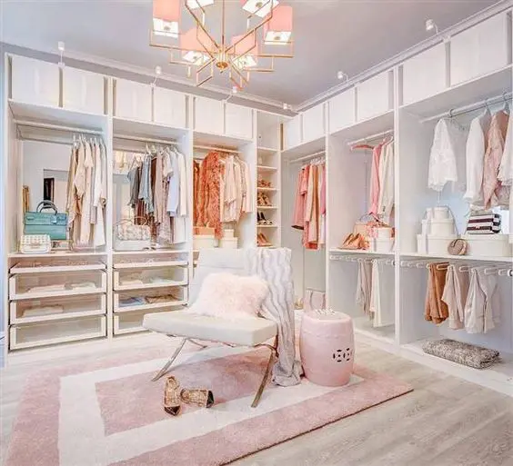 a glam feminine closet with open storage units, drawers, a pink chandelier, a chair and a side table plus a pink printed rug