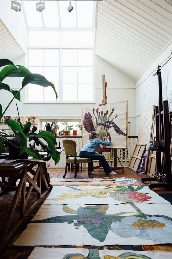 A light filled artist studio with skylights and windows, easels, bookcases, storage shelves, potted plants, colorful artwork