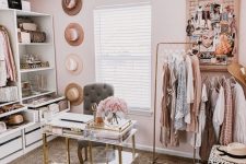 a lovely pink closet with open storage units and drawers, a makeshift clsoet, a crystal chandelier, a mini desk and a grey chair plus hats as decor